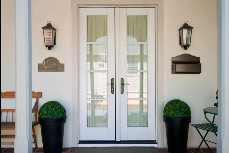 Double white French doors with divided lites, black carriage lanterns either side, boxwood topiary in black planters, and a red quarry-tile porch floor.