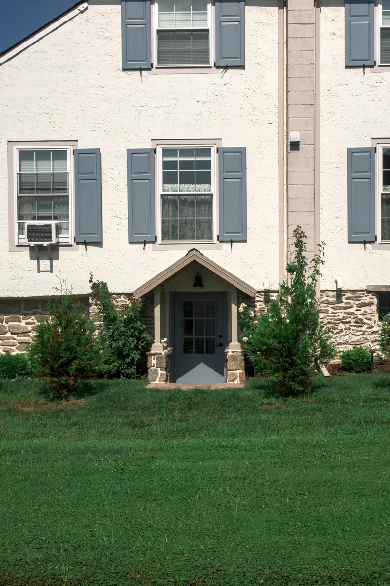 The side door under its little gabled hood, a lantern above it and young trees either side, seen across the lawn.