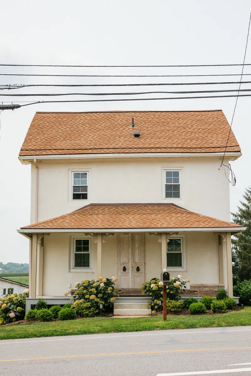 The farmhouse straight on from the road — stucco front, deep porch, and hydrangeas along the foundation.