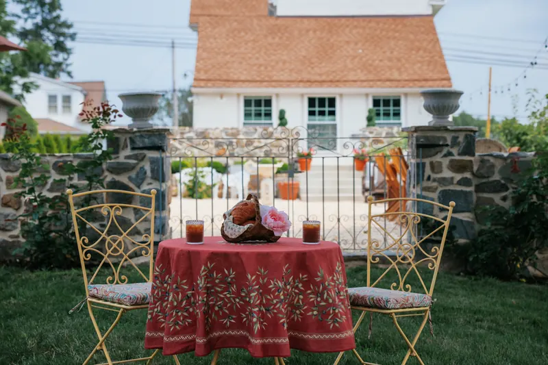 A round garden table in a red Provençal cloth with two gilt bistro chairs, the stone gateposts and the house beyond.