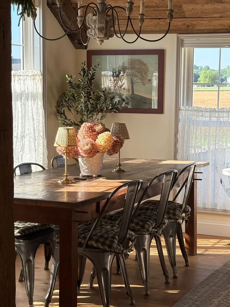 The dining room — the long farm table under a chandelier, with the farmland visible through the window beyond.