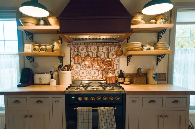 The kitchen — a black range under a timber hood, copper pans on a rail against patterned tile, open shelves of pottery either side.