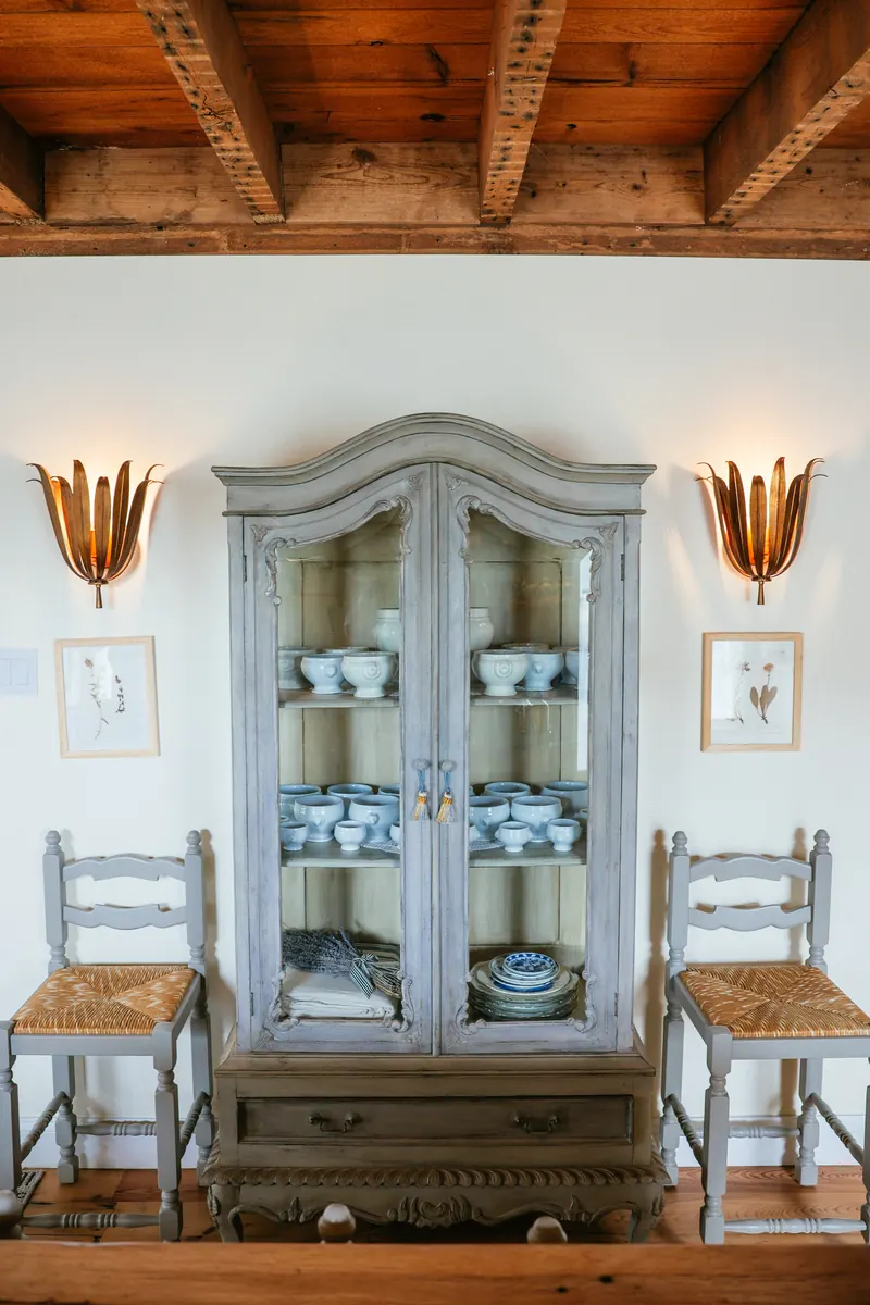 A grey painted vitrine of blue-and-white china between two rush-seat chairs and gilt sconces.