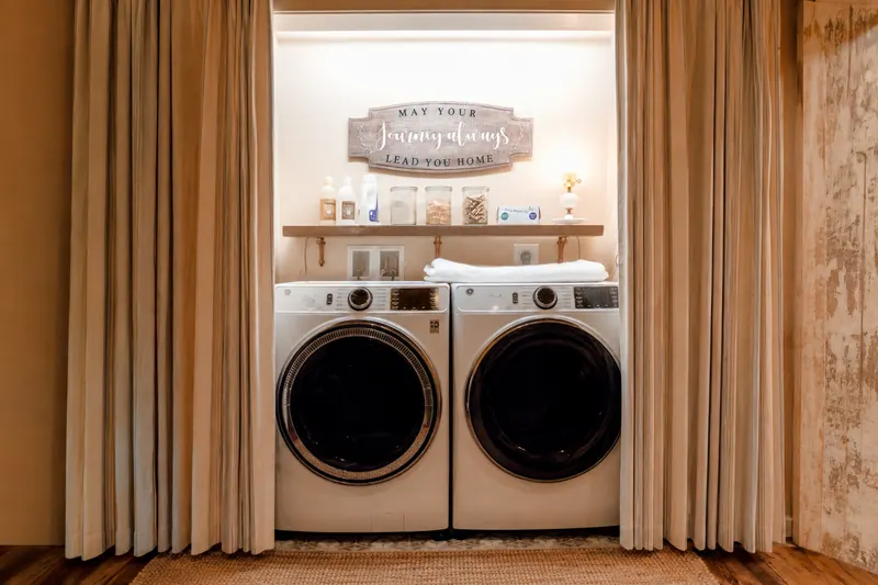 A laundry nook off the hallway — a full-size washer and dryer behind striped linen curtains, under a shelf of glass jars and a wooden sign.